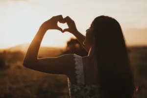 Femme qui fait un coeur avec les mains devant un coucher de soleil